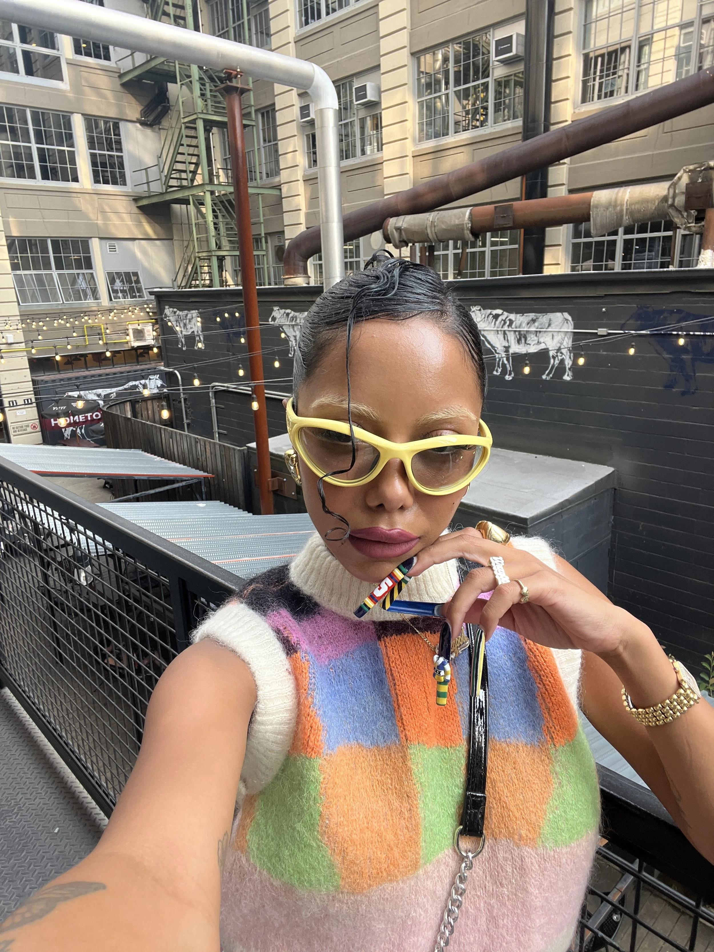 A woman wearing yellow goggles, a multicolored sleeveless sweater, multiple rings, and a gold bracelet, taking a selfie in an urban outdoor setting with industrial pipes, a black fence, and black wall with white animal illustrations in the background