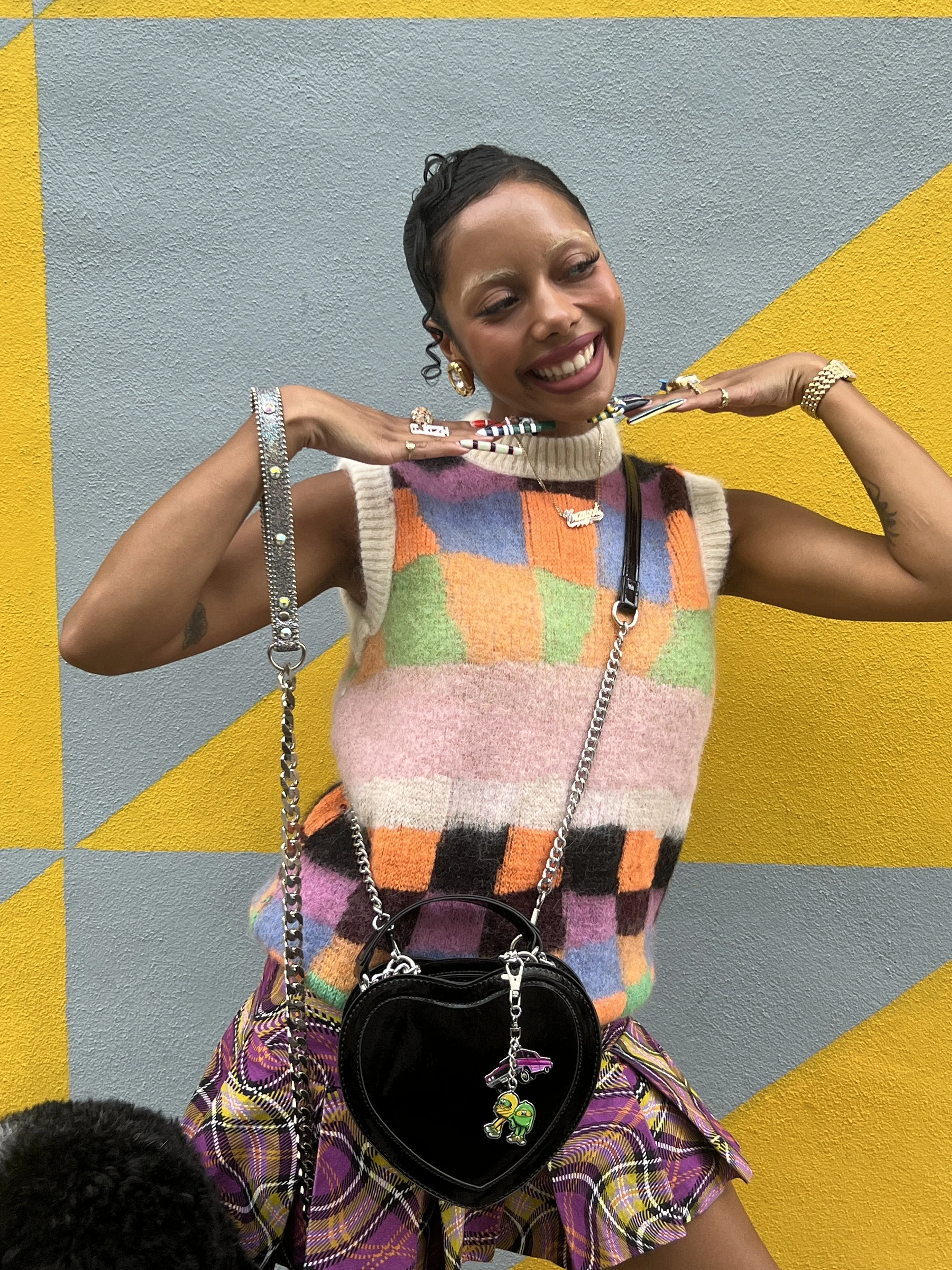 A young woman smiling and posing with her hands near her shoulders against a colorful yellow and gray geometric wall. She is wearing a sleeveless multicolored sweater vest, patterned purple plaid shorts, and has a heart-shaped black purse with colorf