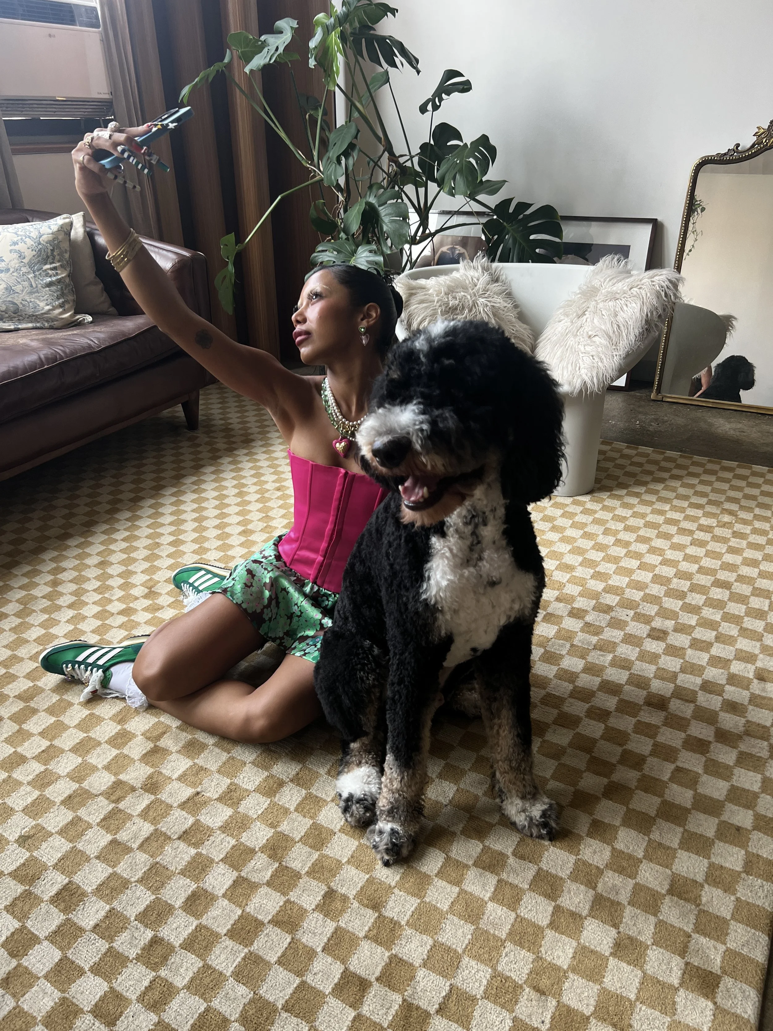 A woman taking a selfie with a large black and white dog sitting in front of her on a checkered beige carpet in a living room. The woman is kneeling on the carpet, holding a phone, wearing a pink strapless top, green patterned shorts, and green sneak
