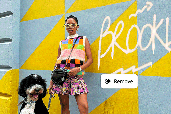 A woman wearing sunglasses, a colorful patchwork top, and a pink plaid skirt poses with a large black and white dog against a vibrant blue and yellow mural wall.