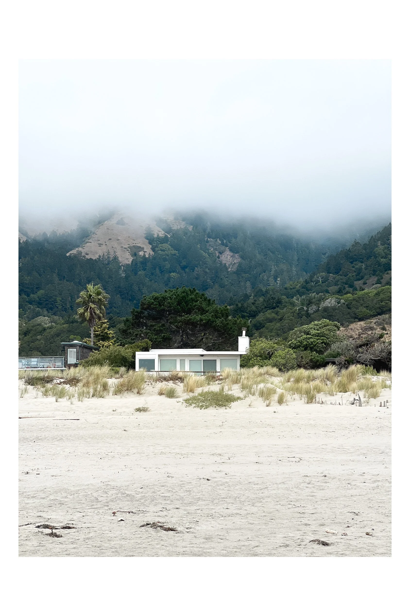 A modern house on a sandy beach, with green vegetation and trees in front of a forested mountain shrouded in clouds. California beach scene.