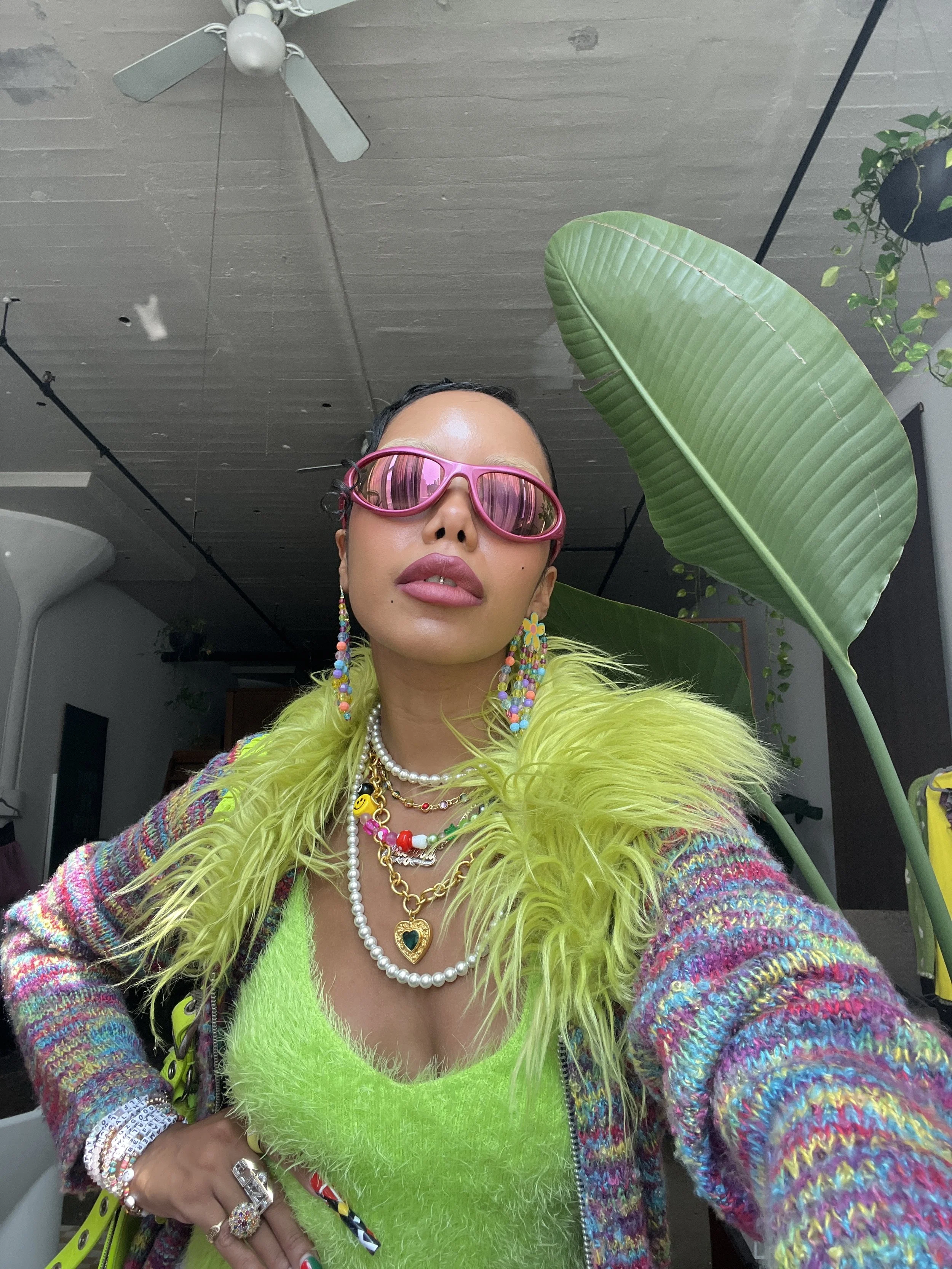 A woman wearing pink sunglasses, layered colorful necklaces, a neon green fuzzy top, and a multicolored jacket with feathered shoulders, standing indoors with large green leaves nearby.