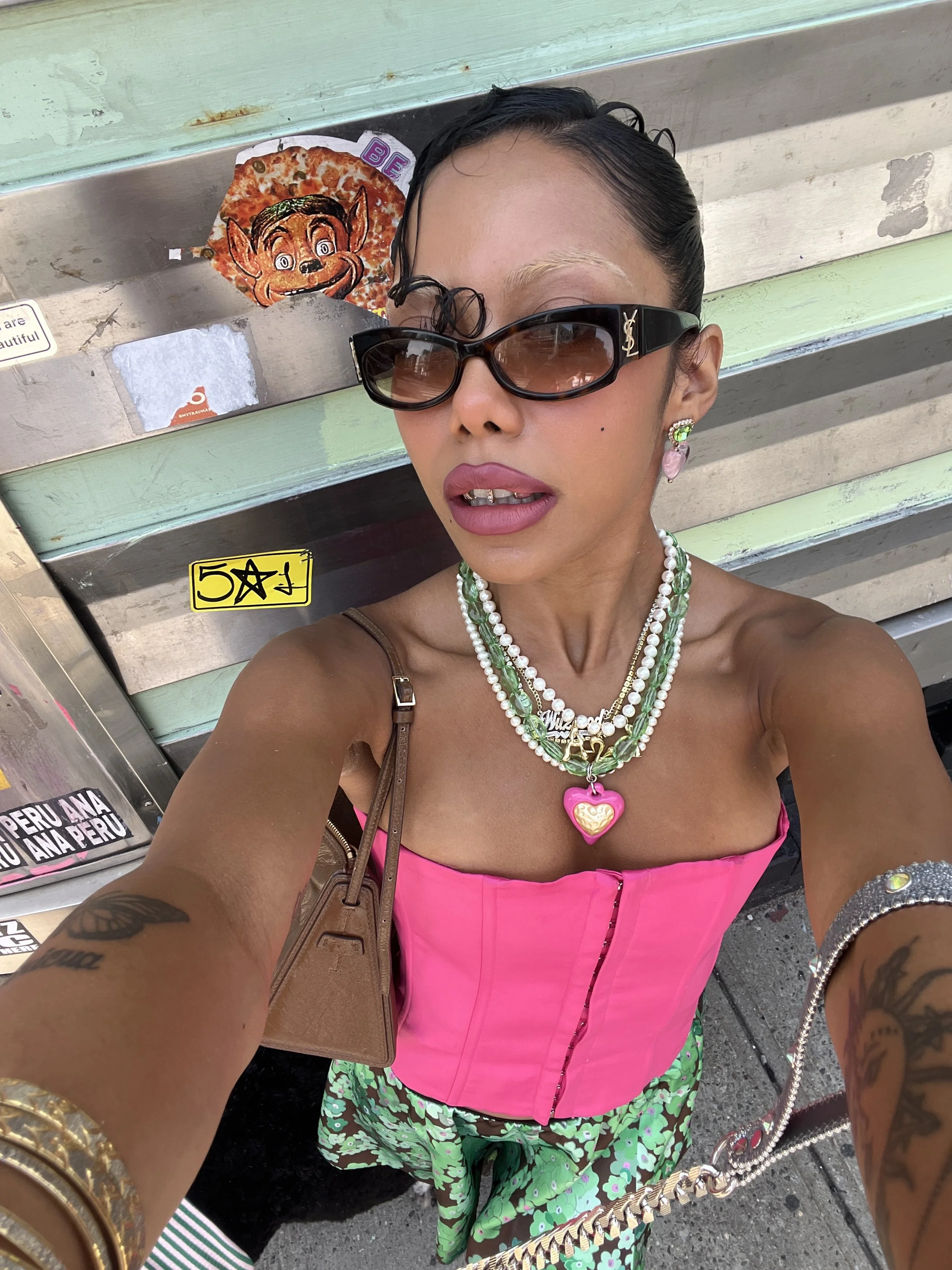 Woman taking a selfie wearing sunglasses, a pink corset-style top, layered jewelry including a heart necklace, earrings, and a bracelet, with tattoos on her arms, standing in front of a metallic surface with stickers and a cartoon sticker of a charac
